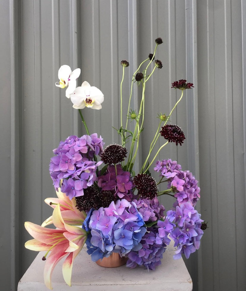 An elegant arrangement featuring vibrant purple hydrangeas, orchids, and lilies, designed by Naked Fern Florals as part of the Helena Soft Hue Floral Arrangement, a luxurious choice for a baby boy floral arrangement perfect for any celebration. #brisbaneflorist #everydayflowers #mothersdayflowers #Nakedfernflowers #weddingflowers